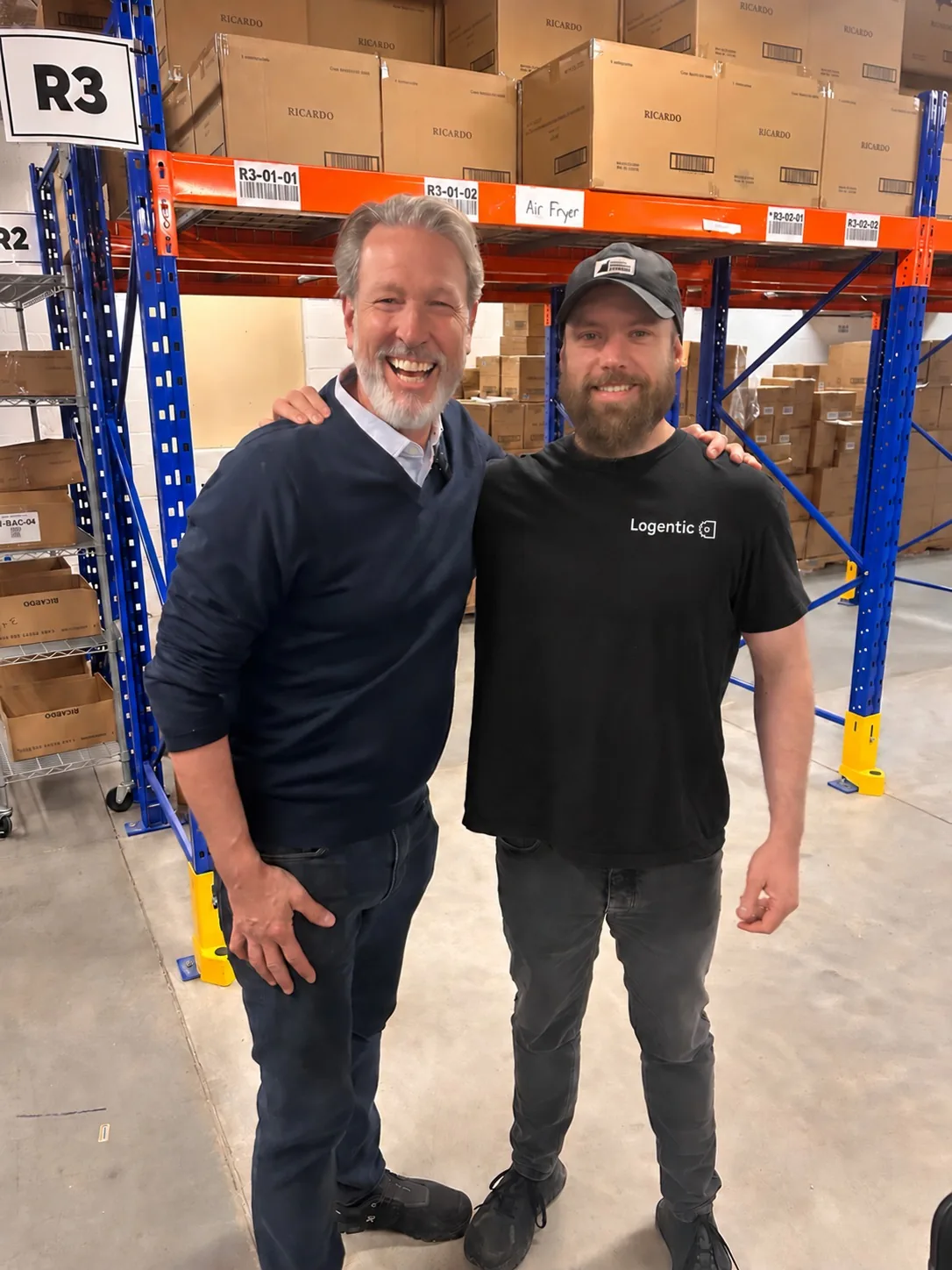 Two people smiling in RICARDO’s warehouse, one in a Logentic t-shirt, in front of racks stacked with RICARDO boxes